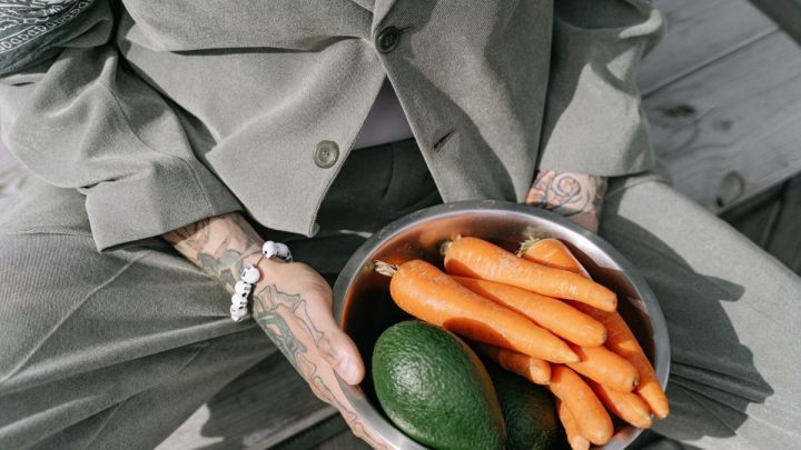 Person in gray suit holding a bowl of fresh carrots and avocados outdoors.